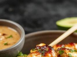 A close-up of skewered grilled chicken satay served on a rustic wooden platter, glazed with a shiny, slightly charred surface. Accompanied by a small bowl of rich, creamy Thai peanut sauce, garnished with chopped peanuts and cilantro. The background features fresh lime wedges and sliced vegetables for a vibrant, appetizing presentation.