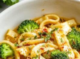 A vibrant plate of broccoli pasta featuring al dente spaghetti topped with bright green broccoli florets, drizzled with olive oil and sprinkled with grated cheese, presented on a rustic white plate with fresh herbs on a wooden table