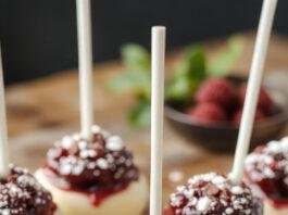 A close-up of enchanting blackberry cheesecake pops arranged on a rustic wooden platter. The pops are coated in a glossy dark chocolate shell with vibrant purple-black blackberry pieces embedded in the creamy white cheesecake center. They are garnished with a sprig of fresh mint and a drizzle of white chocolate, creating a striking contrast. The background features softly blurred forest elements, enhancing the magical forest theme.