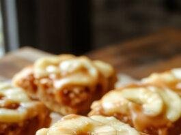 A close-up of golden-brown salted caramel apple bars topped with a drizzle of caramel sauce and sprinkled sea salt. The bars reveal a layered appearance with a crumbly crust, tender apple filling, and glossy caramel topping, all neatly sliced and arranged on a rustic wooden serving board with a few apple slices beside them.
