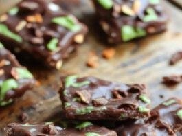 A rectangular piece of dark chocolate bark decorated with white melting chocolate in the shape of a spooky witch silhouette, with candy eyes and orange sprinkles creating a festive Halloween theme, presented on a rustic wooden surface.