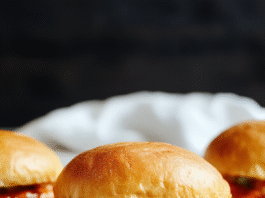 Close-up of mini buffalo chicken sliders on soft brioche buns, glazed with tangy orange-red buffalo sauce, topped with crumbled blue cheese and chopped celery, on a wooden cutting board with a side of ranch dip in a small white bowl.